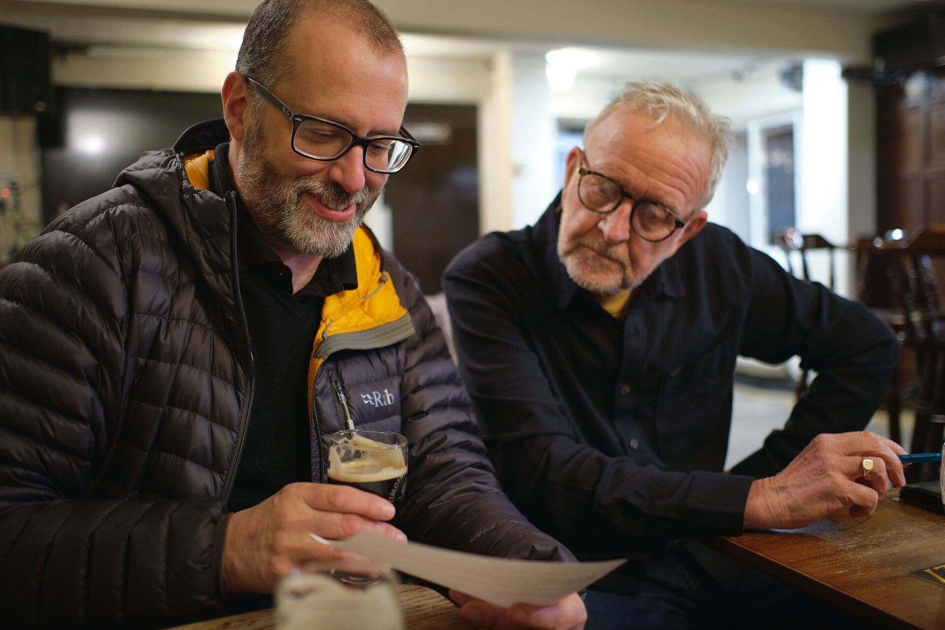 Two older men with short hair and black glasses, holding half a dark beer and looking at a quiz question sheet in the Little Angel, Whitby, at Whitby Hot Chocolate Festival