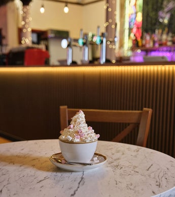 A white cup and saucer piled high with whipped cream and coloured sprinkles, on a white marble table in front of a cocktail bar in Whitby, North Yorkshire