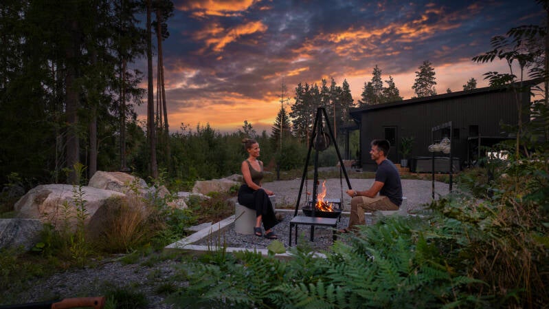 Pariskunta istuu nuotiolla romanttisessa kesäillassa, kun aurinko värjää viimeisillä säteillään pilvet luonnon omaksi taideteokseksi!