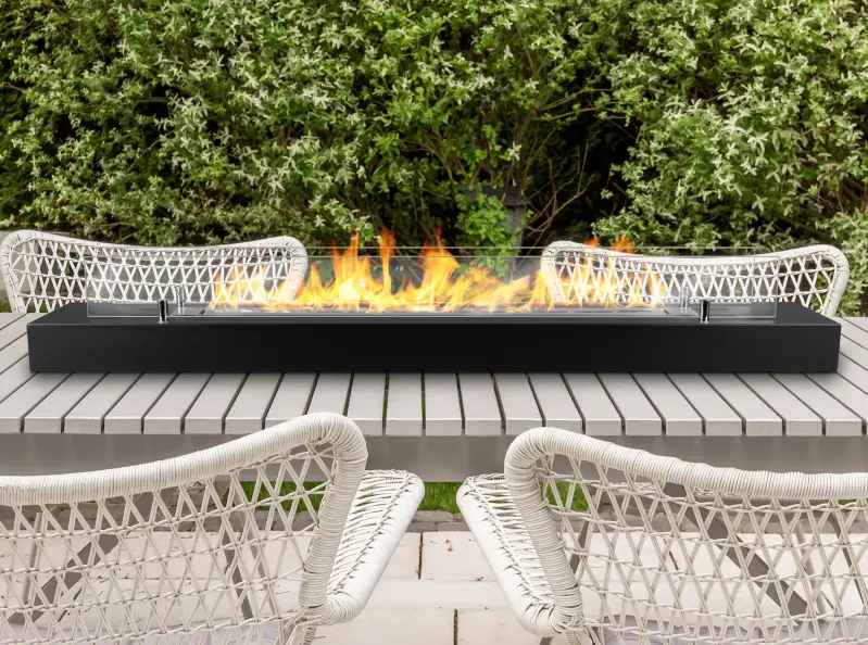 Lange bio-ethanol haard van 1200 mm op een lichtgrijze buitentafel met gevlochten stoelen, omgeven door groene tuinbeplanting