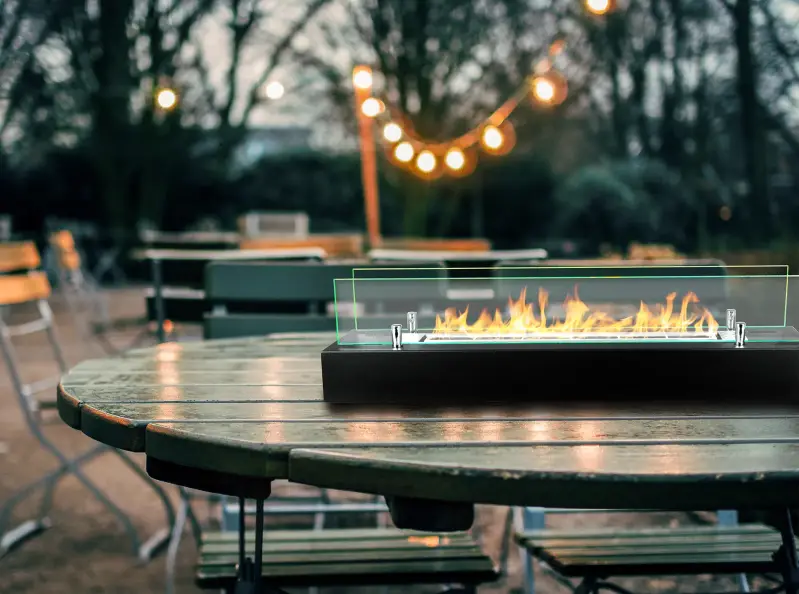 Bio-ethanol haard met glas van 700 mm op een ronde houten buitentafel, omringd door stoelen en zachte verlichting van lantaarns in de achtergrond