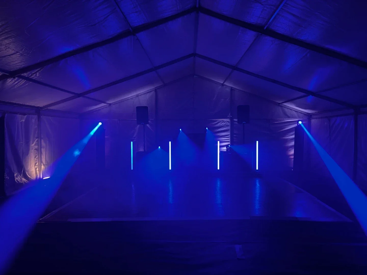 Black MHX DJ setup in a marquee, with blue moving-head beams, speakers on podiums and vertical light bars