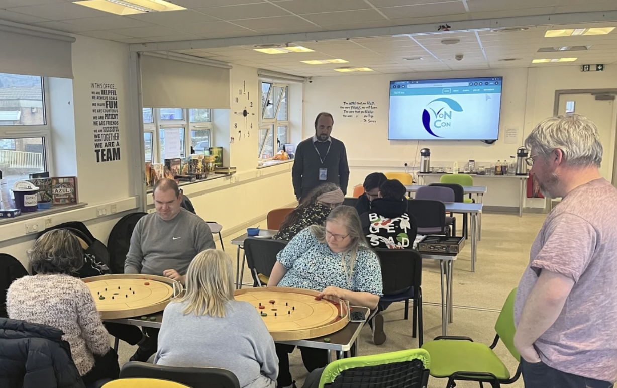 Three tables of 2-3 people playing different board games. There are two people standing up, one in the background, one in the foreground. There is a screen showing the VoNCon logo in the background. 