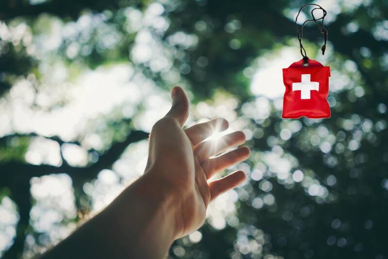 Eine Hand streckt sich im grünen Wald dem Licht entgegen und greift nach einem roten Erste-Hilfe-Kit mit weißem Kreuz