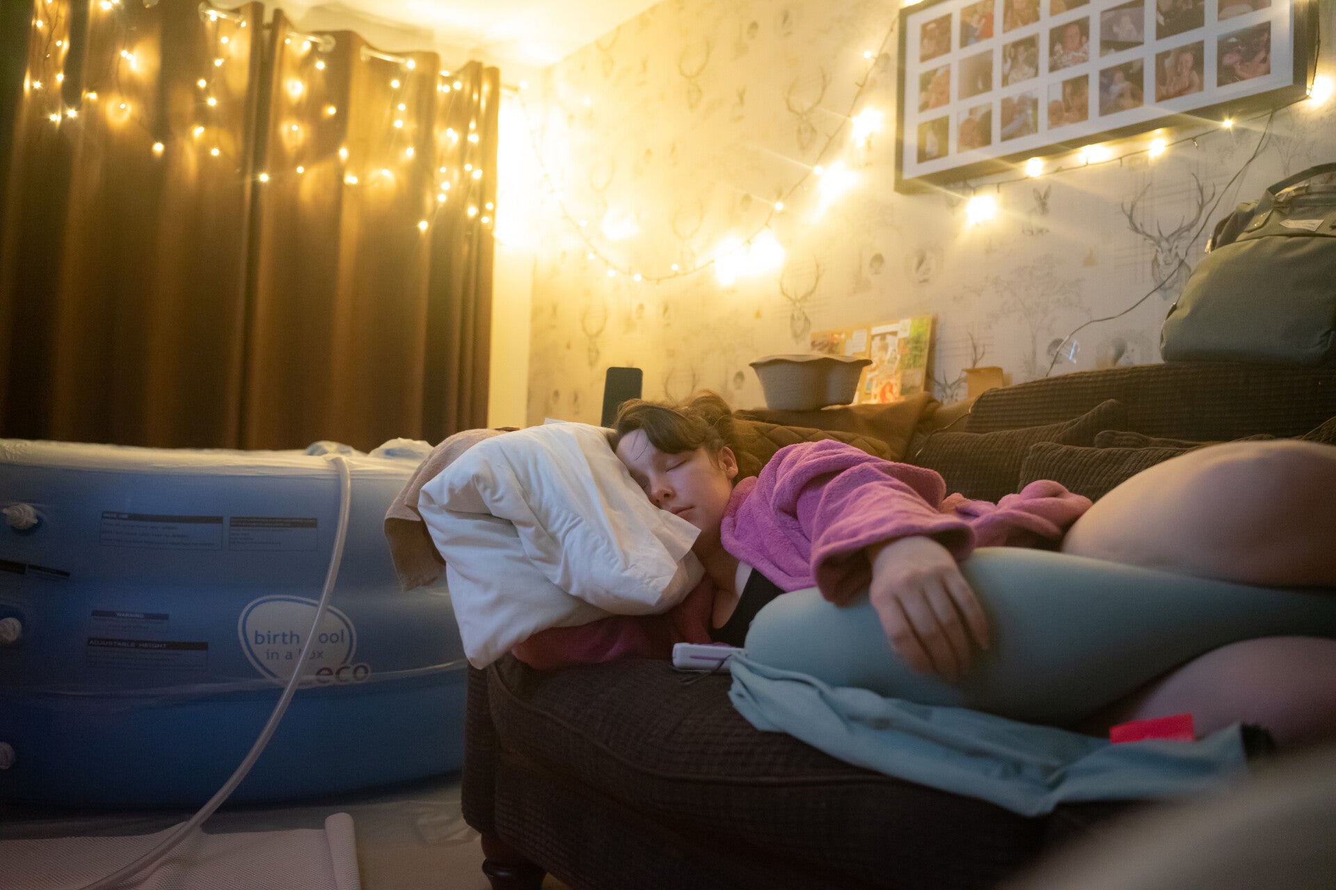colour image of a dark haired woman in labour resting on a sofa at home propped up with pillowsi. The lights are dim, there are fairy lights in the background and a birth pool near . Home Birth in Cheshire. Photo by mamakat.co.uk