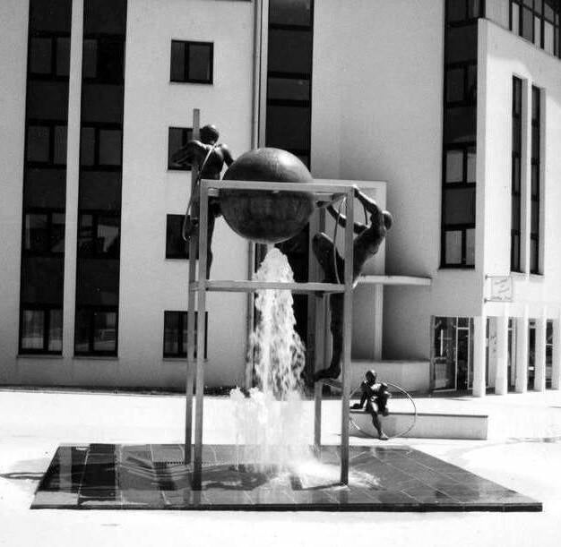 Deux personages en cuivre martelé grimpent sur une structure en inox au sommet de laquelle se trouve une sphère en cuivre martelé semblant portée par le jet d'eau jaillissant du sol. Au second plan, un enfant en cuivre martelé est assis sur un banc. Fonta
