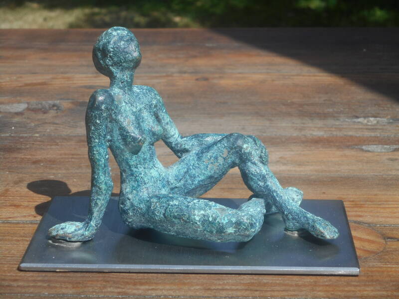 Figurine féminine en bronze assise. Elle est appuyée sur sa main droite, la gauche étant posée sur le genou de la jambe gauche repliée.