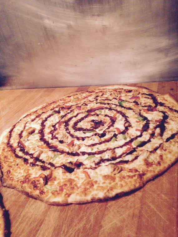 Goldie's Pizza specialty menu configuration: A signature thin-crust artisan pizza featuring a precise, decorative spiral sauce drizzle on a wood service station."