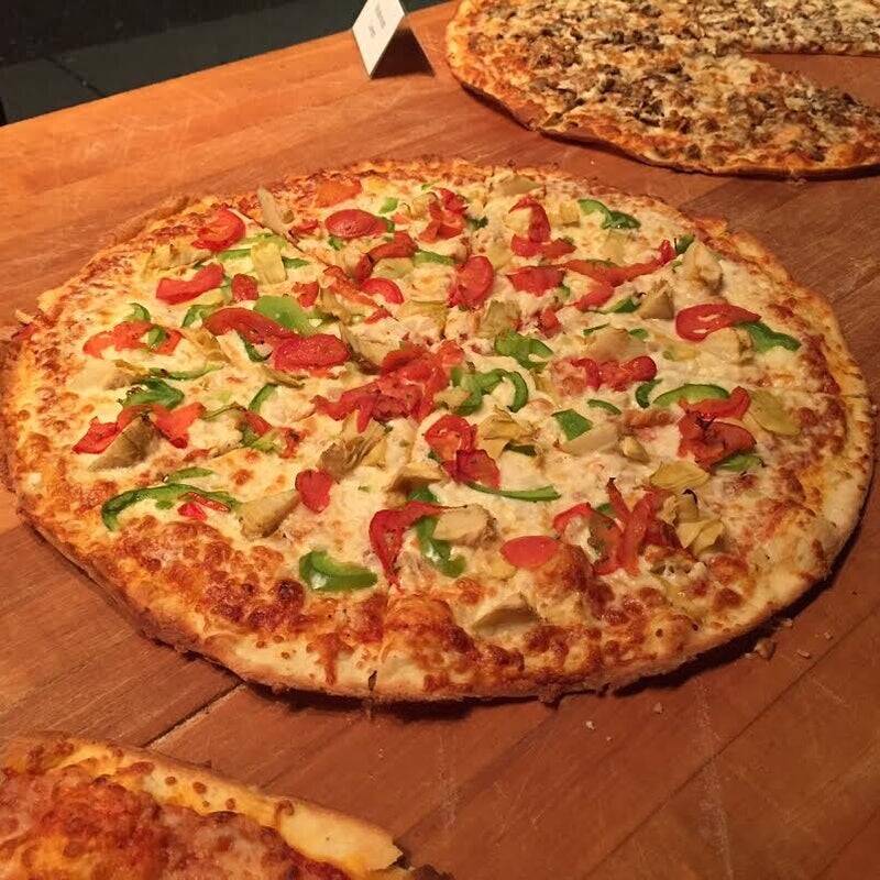 "Goldie's Pizza proprietary recipe presentation: A large artisan vegetarian pizza loaded with vibrant bell peppers, diced tomatoes, and artichoke hearts on a seasoned wood cutting surface."