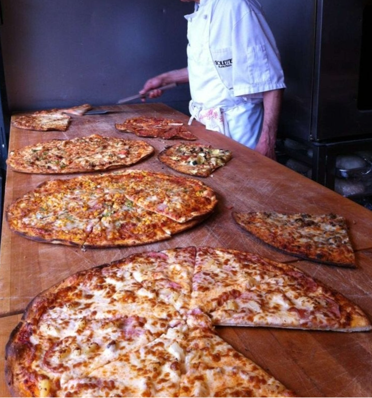 "Goldie's Pizza operational history: An artisan pizza chef slicing a variety of thin-crust pizzas on a massive wood preparation table next to a commercial baking station.