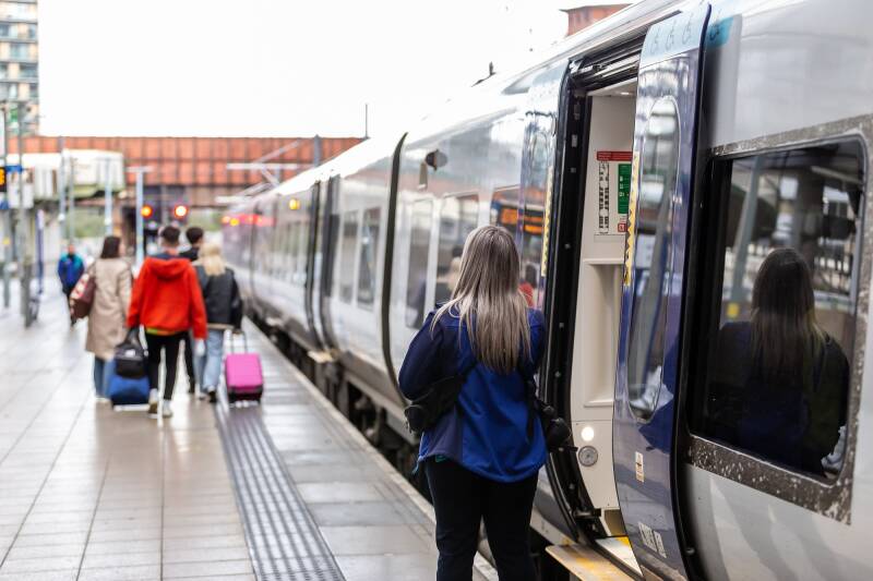 Passengers with luggage walking along a station platform as a commuter boards an open door of a regional passenger train.