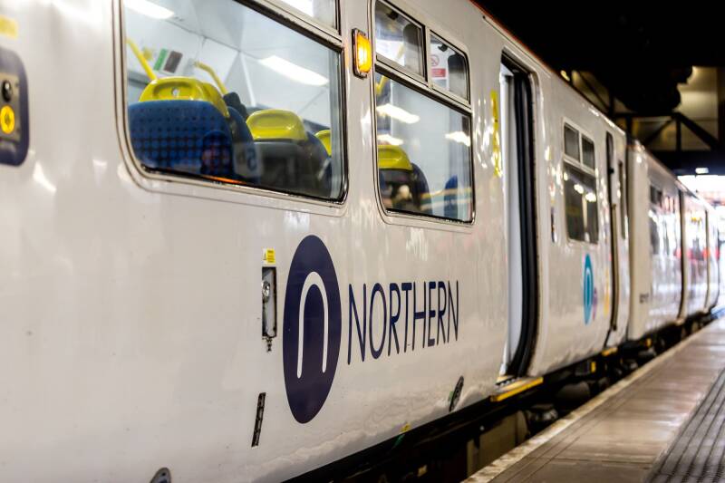 A white Northern Rail passenger train stationary alongside a station platform, highlighting the prominent dark blue Northern branding logo on its side panel.