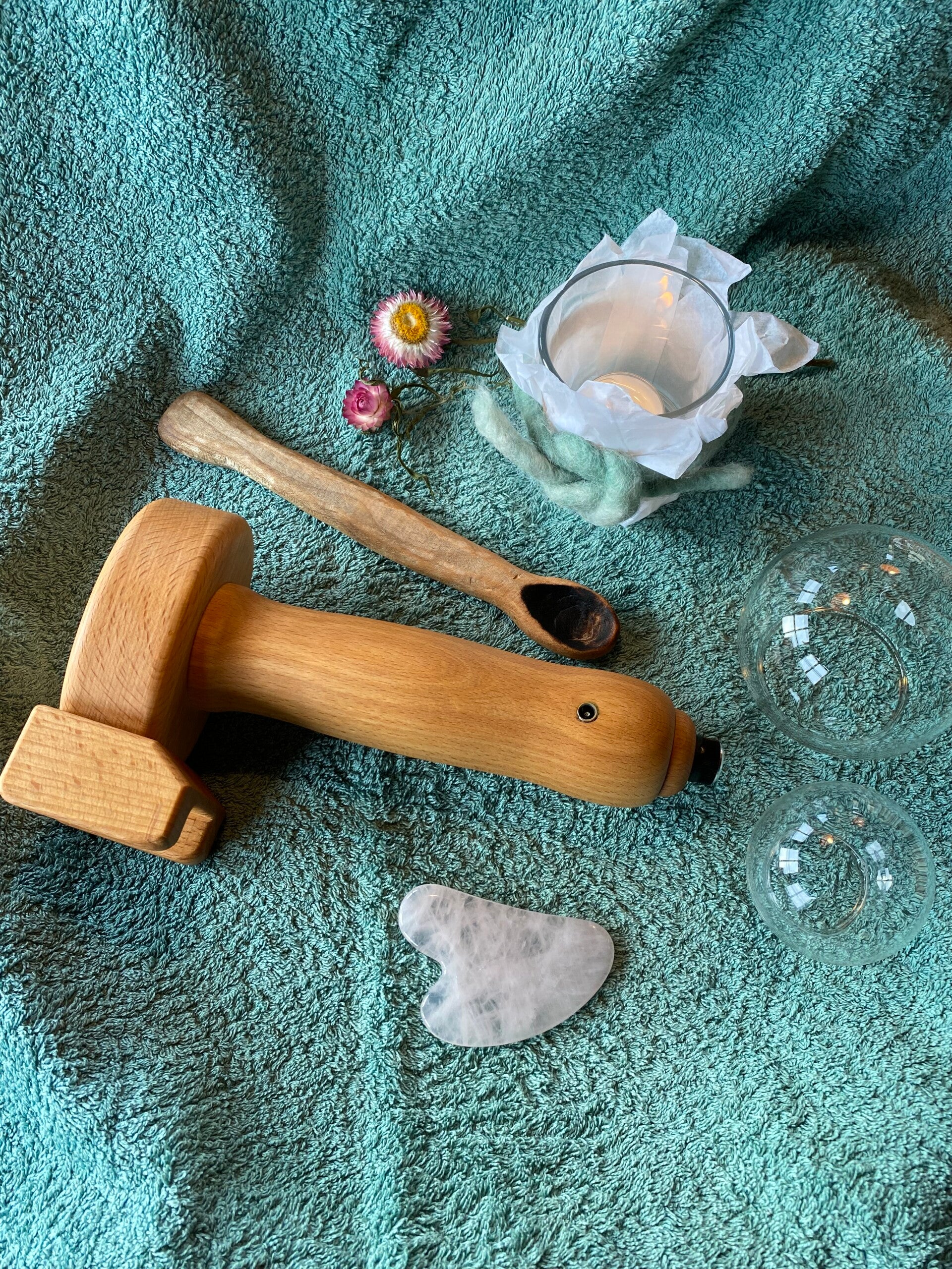 Verschiedene Hilfswerkzeuge der Körperarbeit auf einem grünen Tuch. Darunter ein elektrisches Massage-Gerät aus Holz, ein hölzerner Löffel, ein Gua Sha Stein, Schröpfgläser und zur Dekoration ein Kerze und Blumen. 
