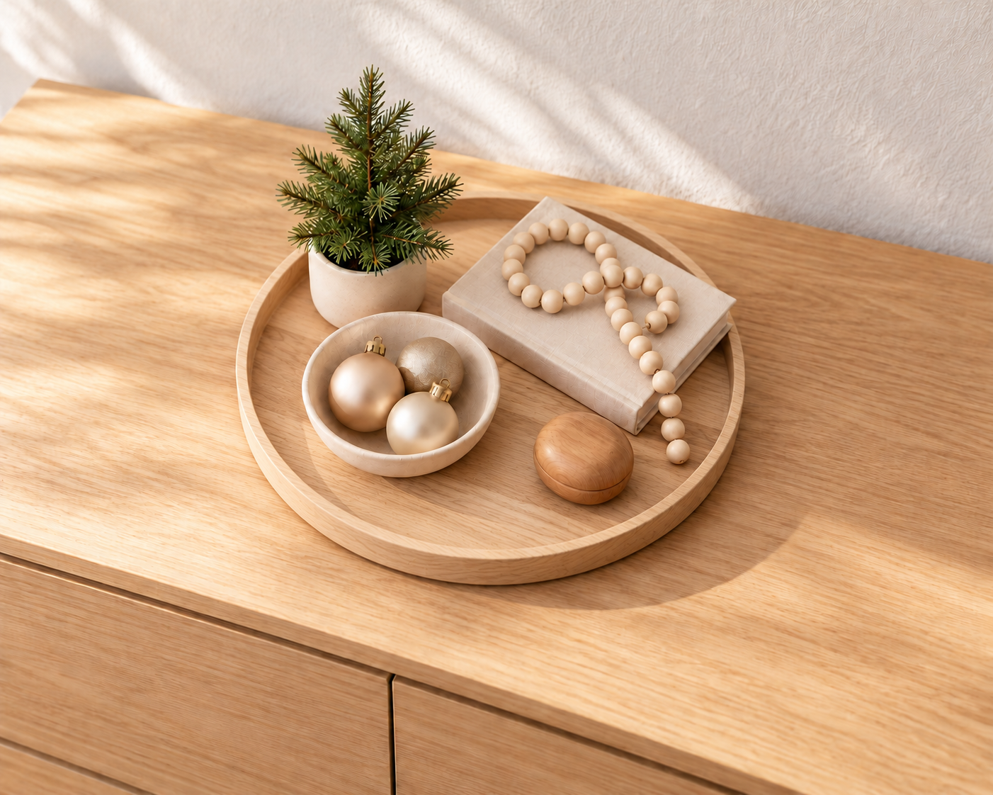 Helle Holzkommode mit terrakottafarbenem Tablett, kleinem Tannenbäumchen, cremefarbenem Dekobuch mit Holzkette, Keramikschale mit drei matten Weihnachtskugeln und Holzdekoration.