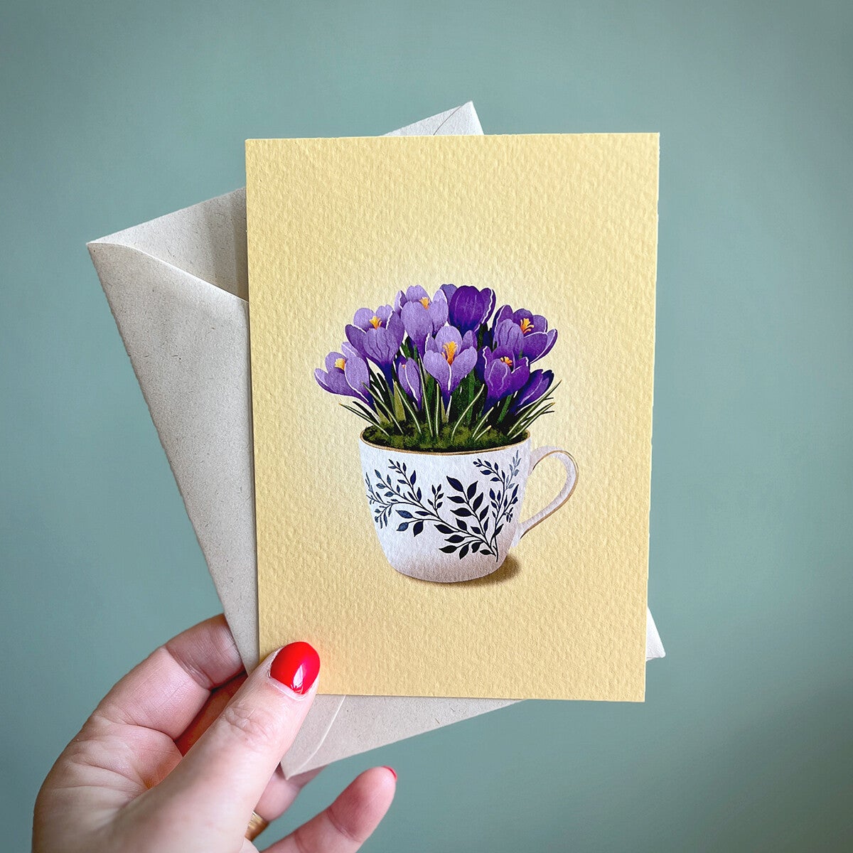 Wenskaart met krokussen die in een mok gepot is. Op de mok staan takjes met blaadjes in een Delfts Blauw motief. De achtergrond van de kaart is zacht geel.