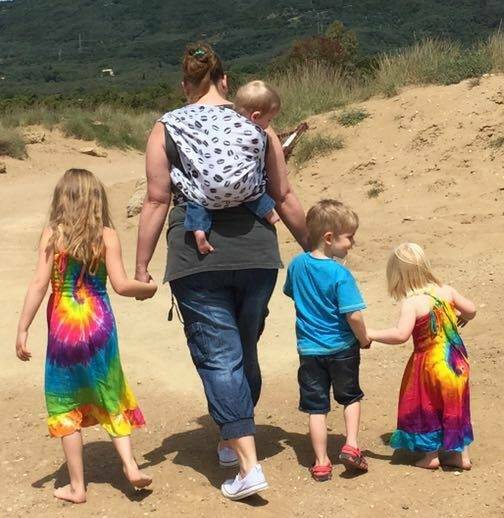 Moeder met zwart shirt die haar kindje op de rug draagt met de Ruck knoop in een Pinkova Opposite Kiss doek. Aan haar linker hand haar dochter in een regenboogjurk. Aan haar rechterhand haar zoon met een blauw shirt. Haar zoon heeft de hand vast van een 2