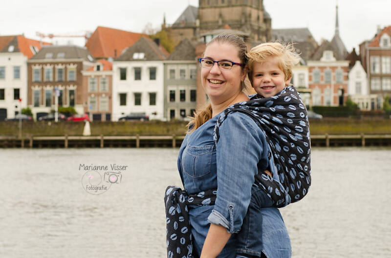 Moeder met zwart shirt die haar kindje op de rug draagt met de Ruck knoop in een Pinkova Blue Moon Kiss.