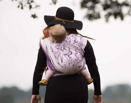 Dubble Hammoch dragen. Moeder met een zwarte hoed die je op de rug kijkt. Moeder heeft een klein kindje op de rug in een draagdoek. De draagdoek is wit met lila van kleur. Het kindje heeft blote voetjes. Op de achtergrond zie je heide en bomen.
