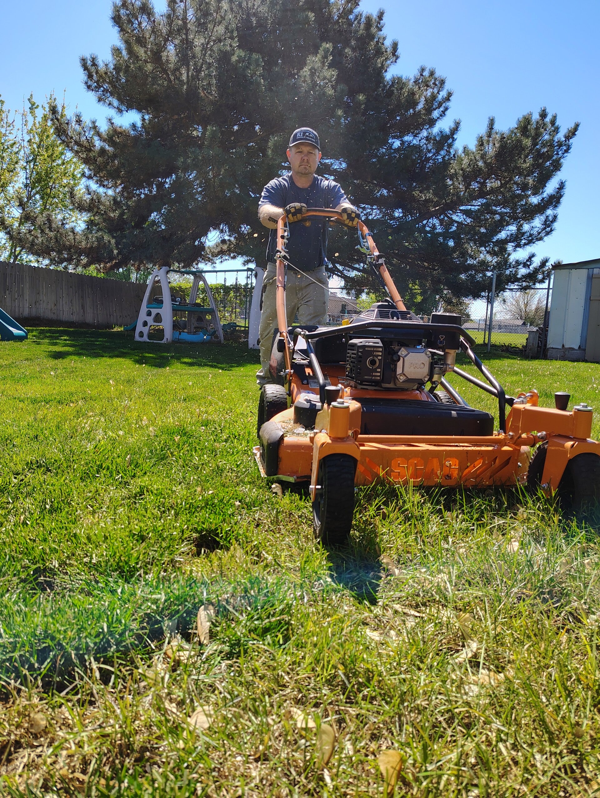 Richard mowing with commerical scag mower coming toward the camera with a green yard and tree in the background