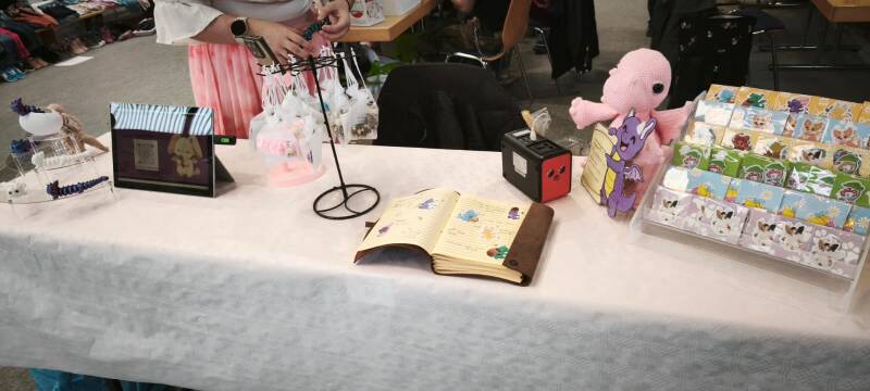 Kleiner Flohmarktstand mit handgemachten Drachen-Produkten, darunter Sticker, gehäkelte Figuren und Sorgenwürmchen im Beutel, liebevoll auf einem Tisch arrangiert, mit Display, Notizbuch und Dekoration
