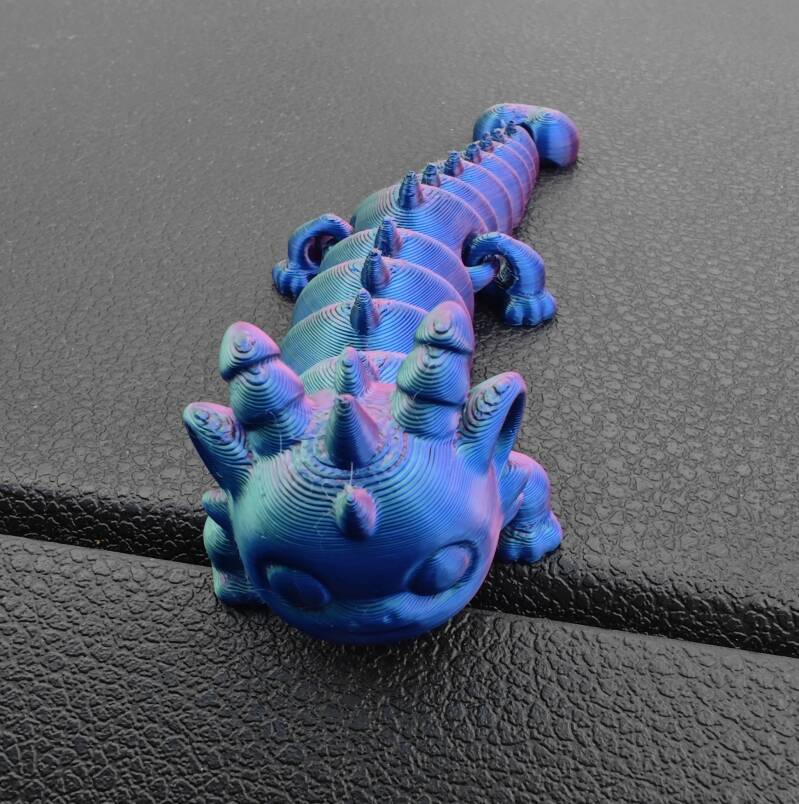 3D-gedruckter, beweglicher Drachen-Flexi in schimmerndem Farbverlauf aus Blau, Lila und Grün, liegend auf einer dunklen Oberfläche, mit segmentiertem Körper und kleinen Hörnern