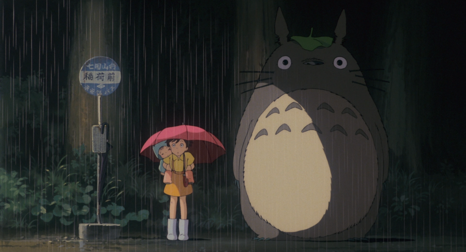Regenszene aus einem Anime: Ein Mädchen mit rotem Schirm wartet an einer Bushaltestelle, neben ihr steht die große Figur Totoro im Regen.