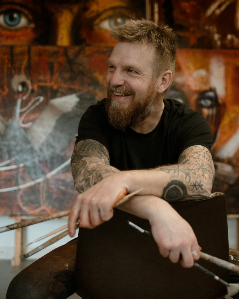 Willem Vos seated in his studio, smiling in front of his large expressive paintings.