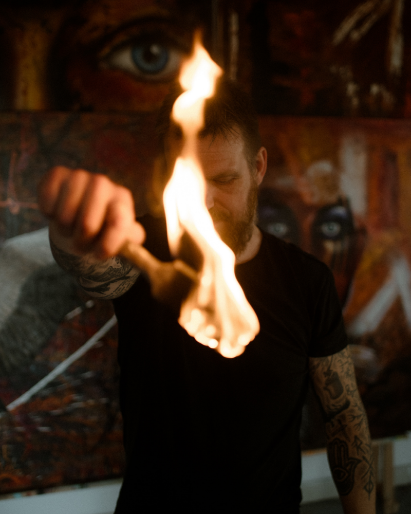Willem Vos standing behind a flame in his studio, surrounded by his paintings.