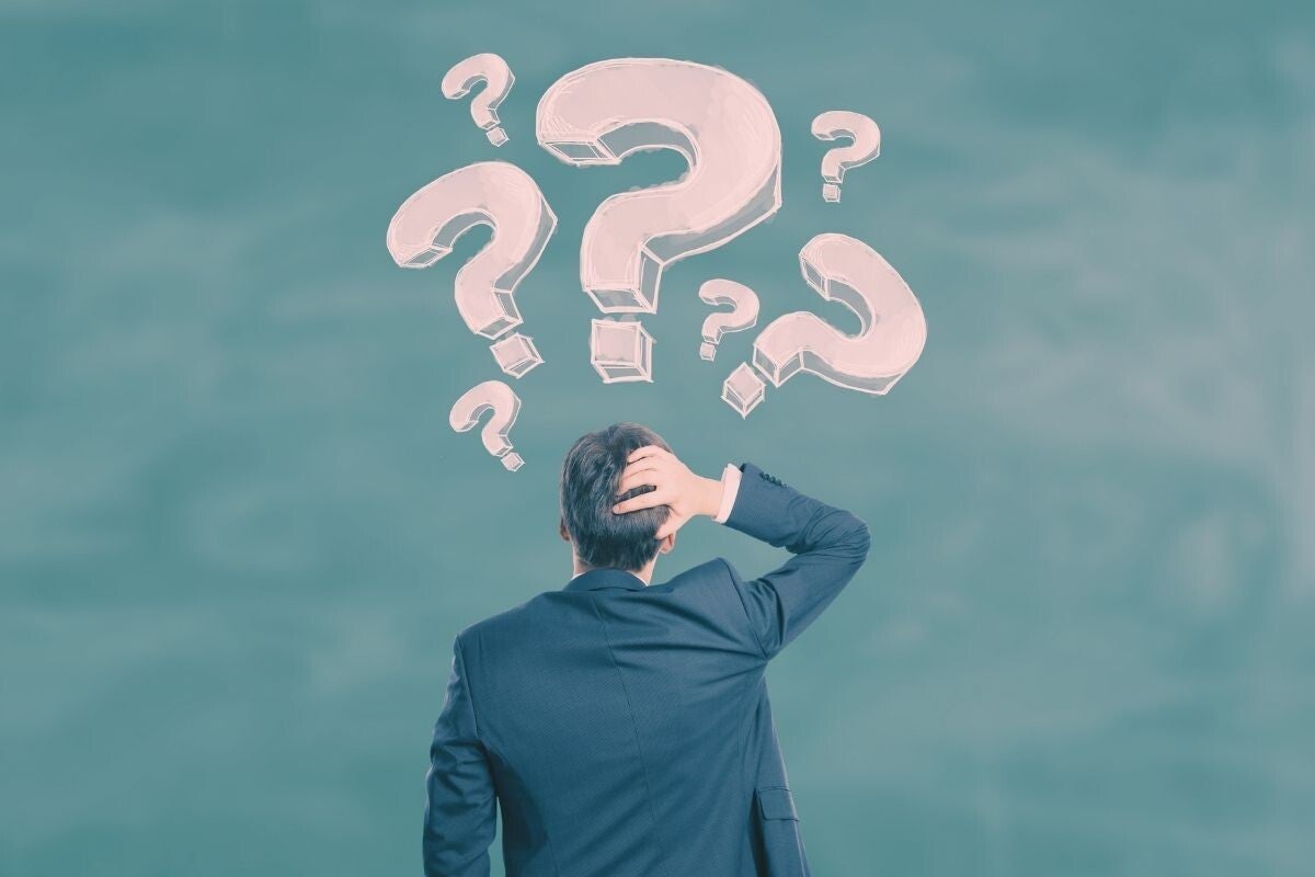 A man in a suit scratching his head while looking up at large question marks in the air.
