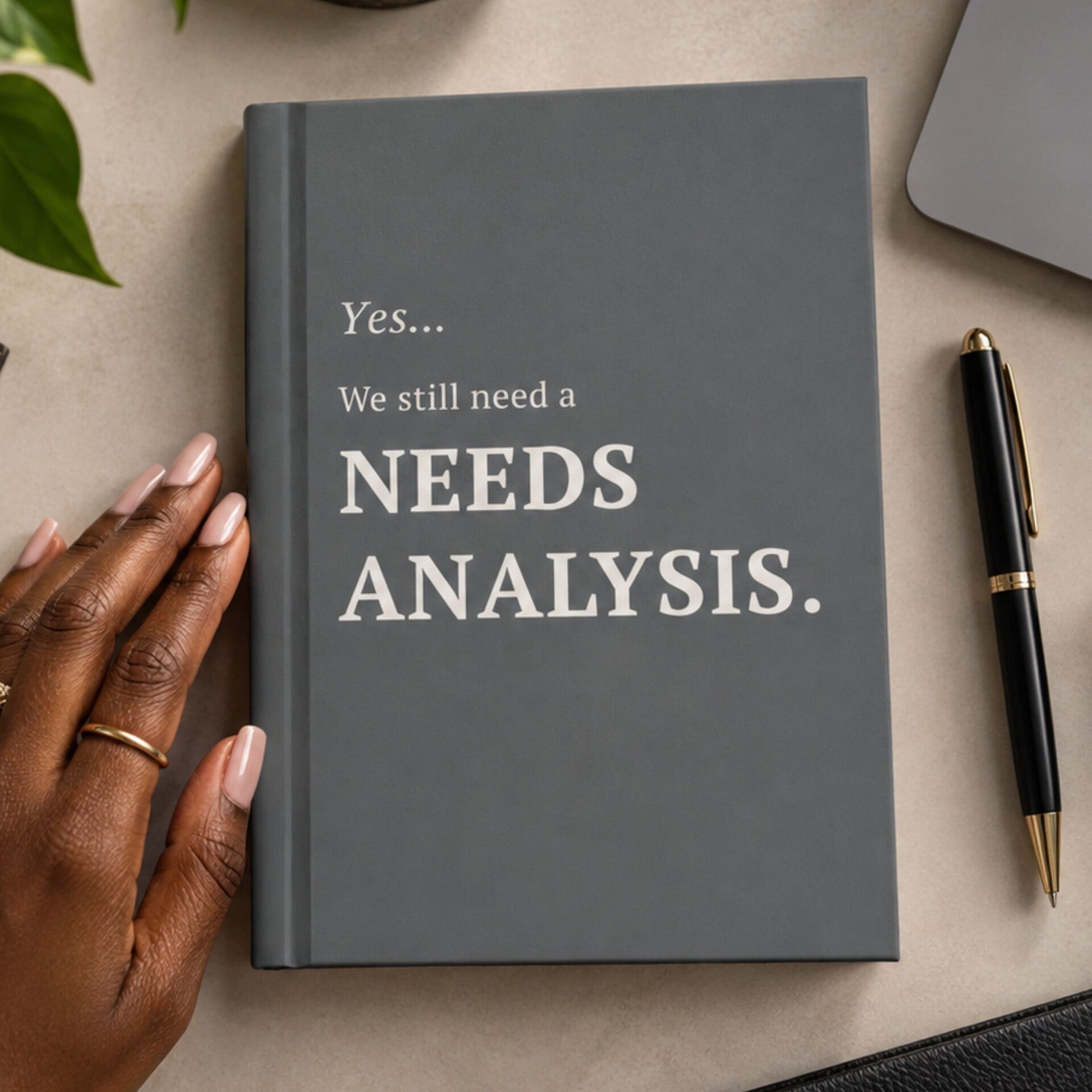 Gray hardcover journal with text in a beige that reads, "Yes...We still need a needs analysis." The journal is on a cream colored desk with a pen on the right and a woman's hand on the left.