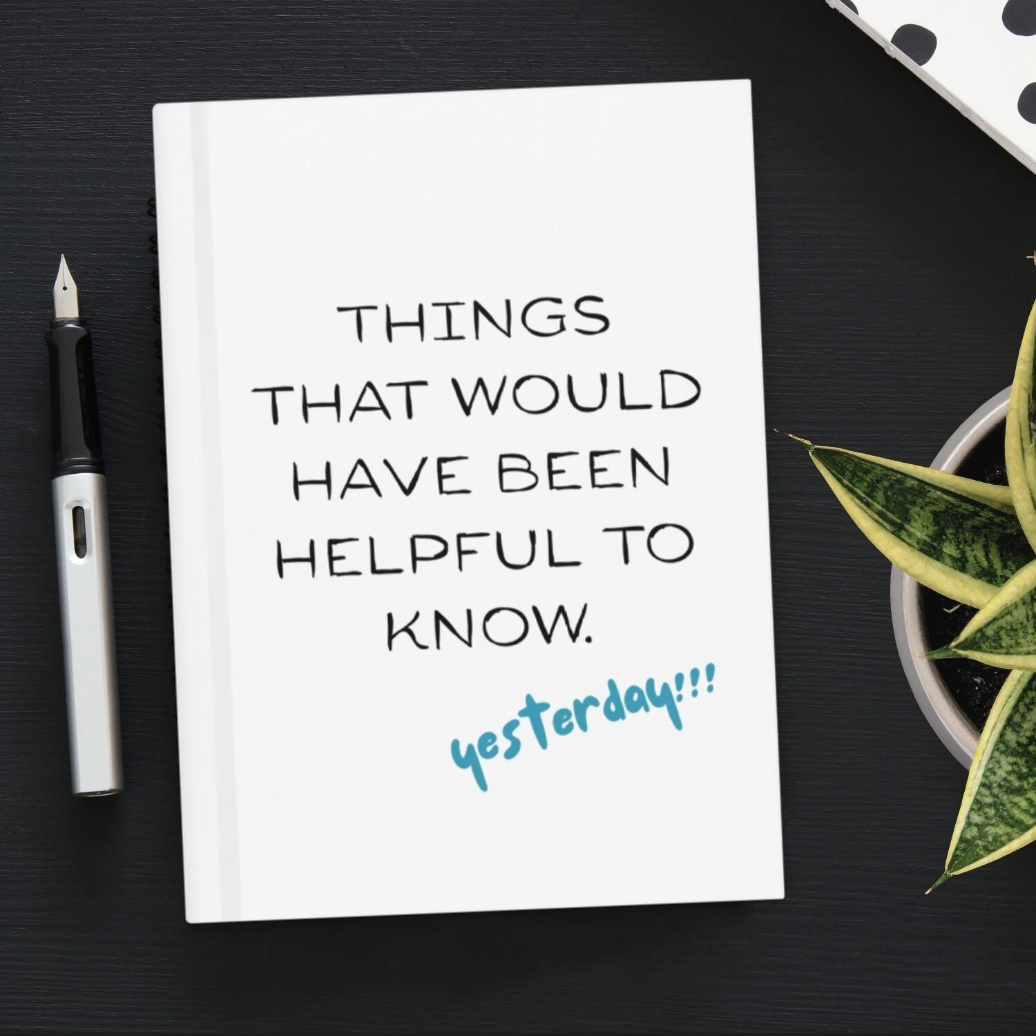 White colored journal with text that reads "Things that would have been helpful to know. Yesterday!!!" The journal is on a black desk with a pen on the right and a plant on the left.