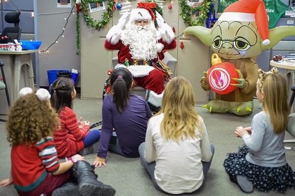 Santa is seated next to Christmas, surronded by gleeful children.