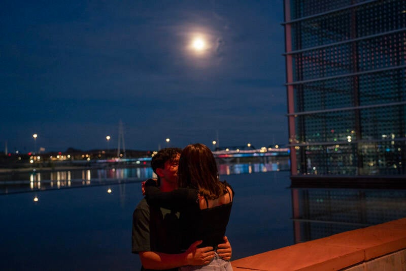 Night time photo with couple holding eachother with the moon and lake in the background. 
