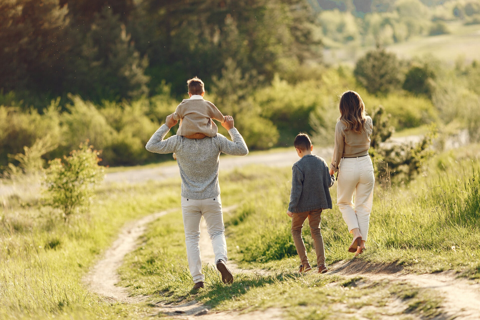 Eine junge Familie geht in der Natur auf einem Rasenweg in Richtung eines kleinen Waldes spazieren. Der Vater trägt ein kleines Kind auf den Schultern, neben der Mutter läuft der ältere Sohn. 