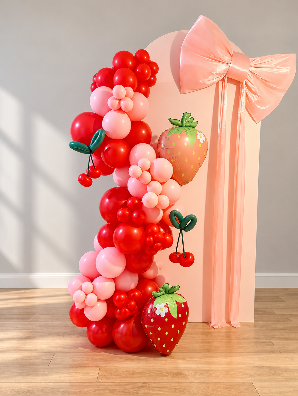 Strawberry and cherry themed organic balloon garland in red and pink beside a peach arch backdrop with a satin bow