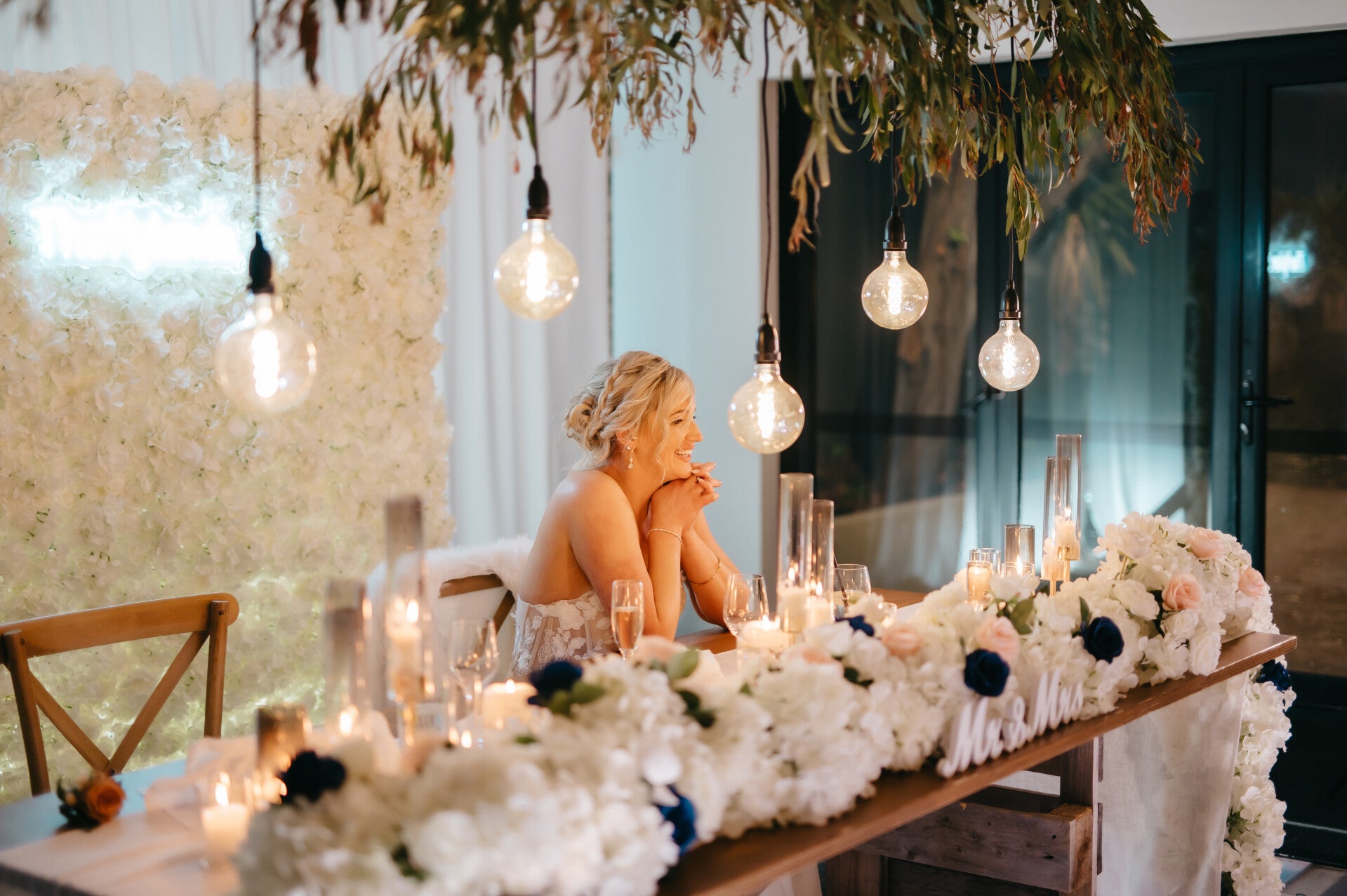 Bride at a sweetheart table styled with a flower wall backdrop and hanging greenery