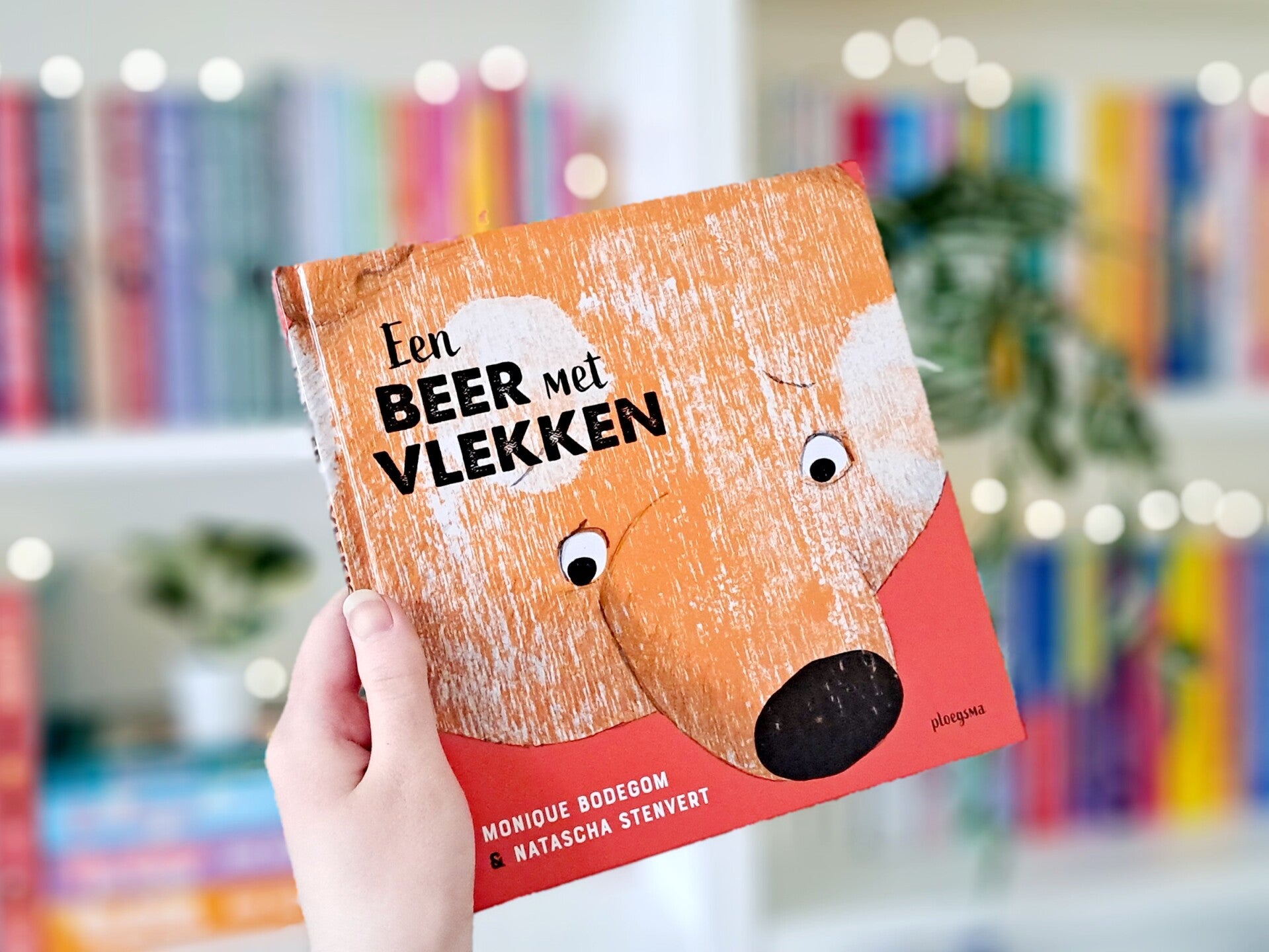 Een witte hand houdt 'Een beer met vlekken'van Monique Bodegom en Natascha Stenvert omhoog voor een kleurrijke wazige boekenkast met lichtjes. Foto bij recensie van turnthepages.nl rolstoelgebruiker en disabled bookstagrammer en blogger