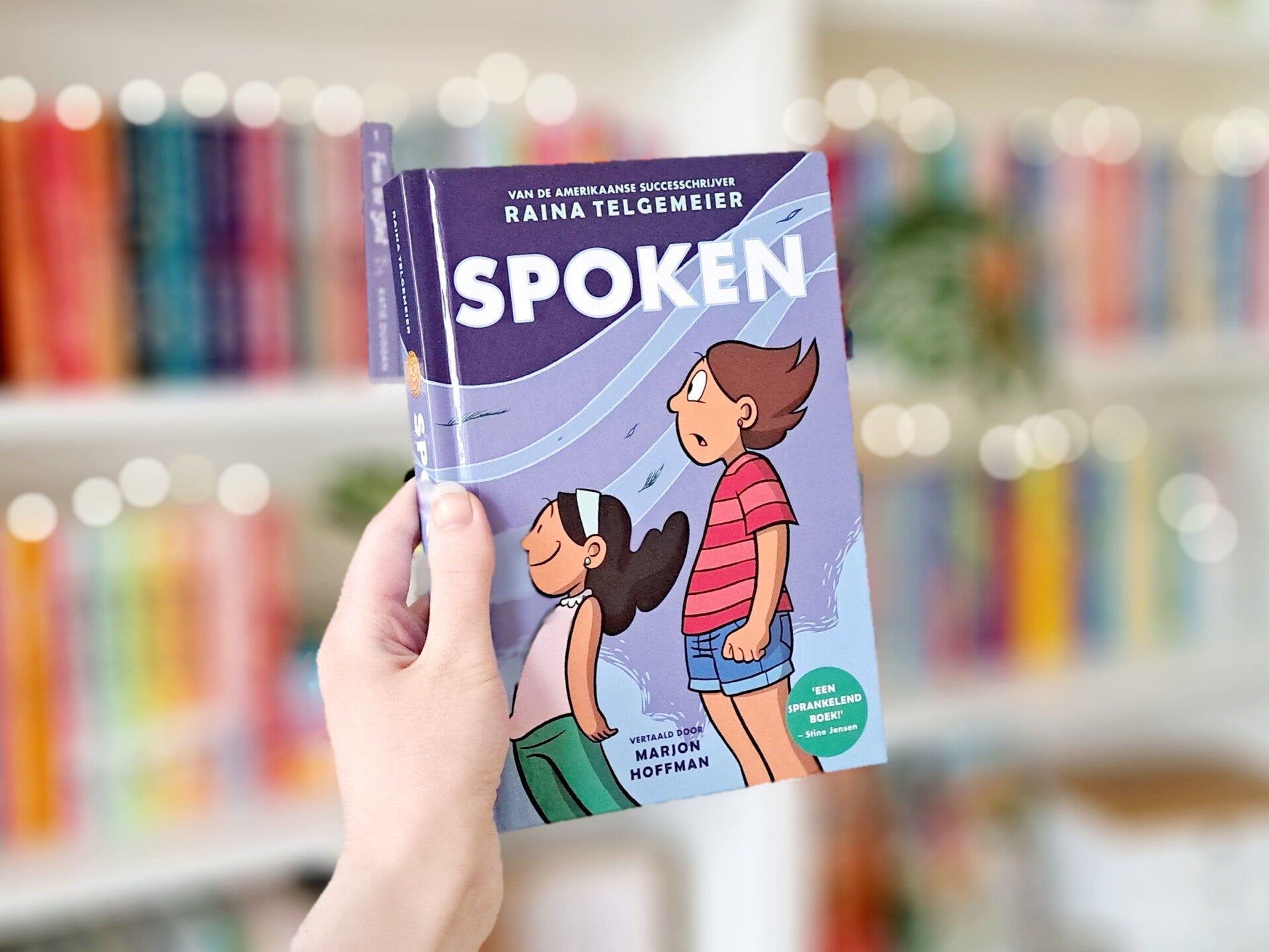 Een witte hand houdt 'Spoken' van Raina Telgemeier omhoog voor een kleurrijke wazige boekenkast met lichtjes. Foto bij recensie door turnthepages.nl Rolstoelgebruiker en disabled bookstagrammer en blogger.