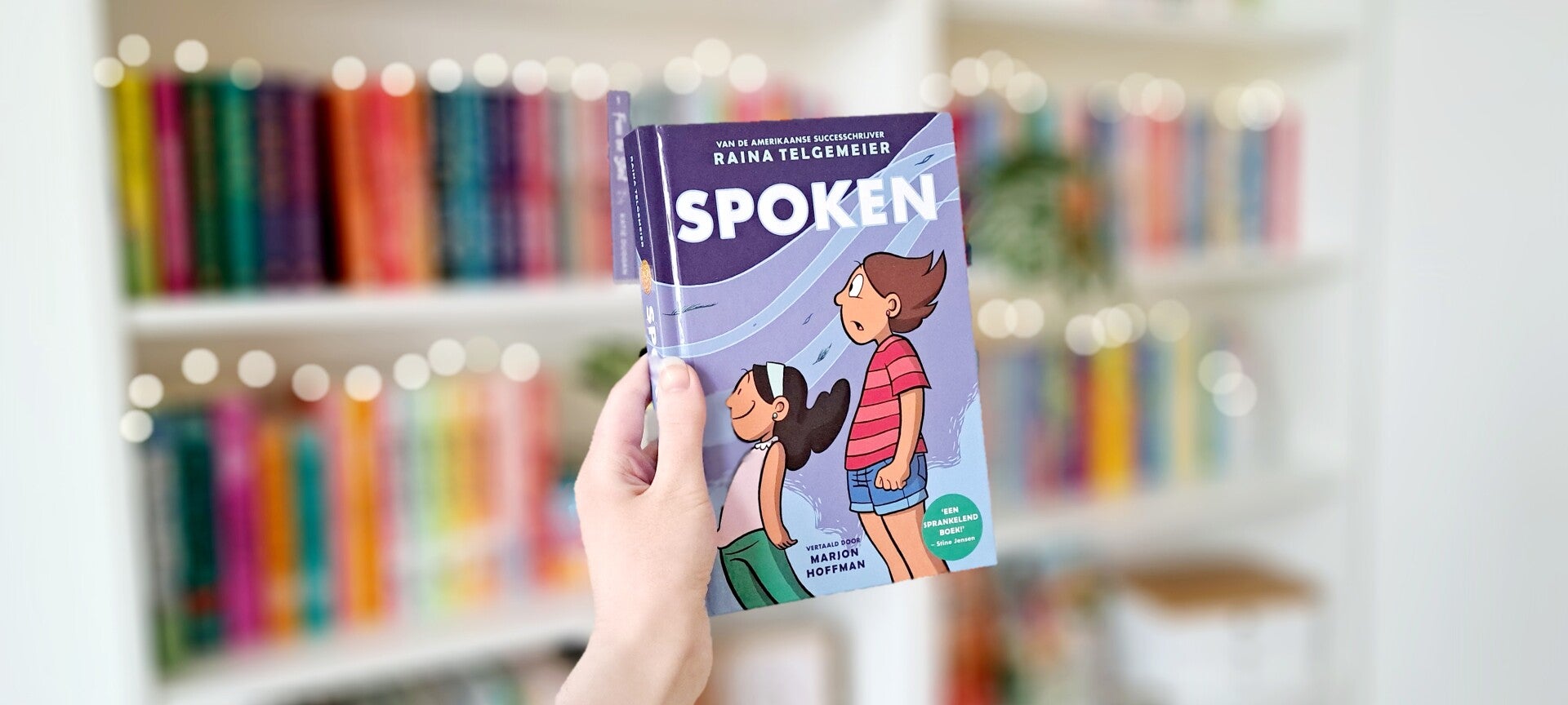 Een witte hand houdt 'Spoken' van Raina Telgemeier omhoog voor een kleurrijke wazige boekenkast met lichtjes. Foto bij recensie door turnthepages.nl Rolstoelgebruiker en disabled bookstagrammer en blogger.