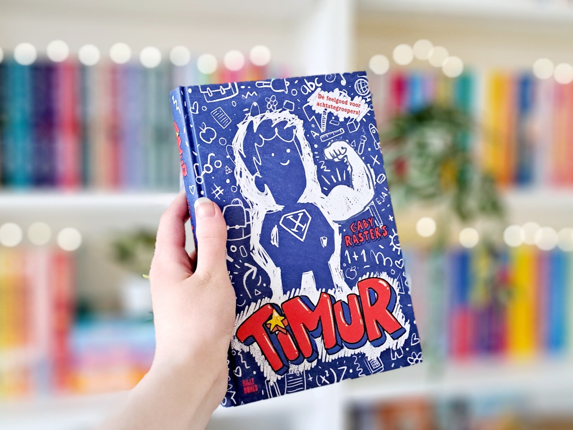 Een witte hand houdt 'Timur' van gaby Rasters omhoog voor een kleurrijke wazige boekenkast met lichtjes. Foto bij recensie door turnthepages.nl Rolstoelgebruiker en disabled bookstagrammer en blogger.