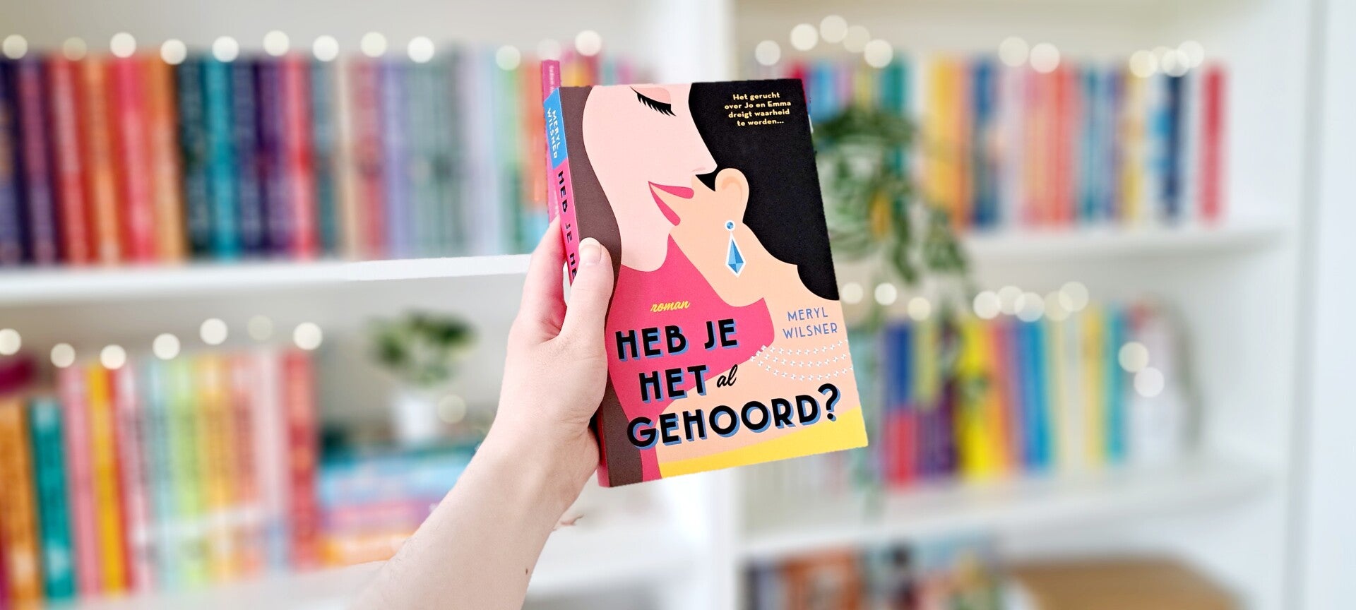 Een witte hand houdt 'Heb je het al gehoord?' van Meryl Wilsner omhoog voor een kleurrijke wazige boekenkast met lichtjes. Foto bij recensie door turnthepages.nl Rolstoelebruiker en disabled bookstagrammer en blogger.