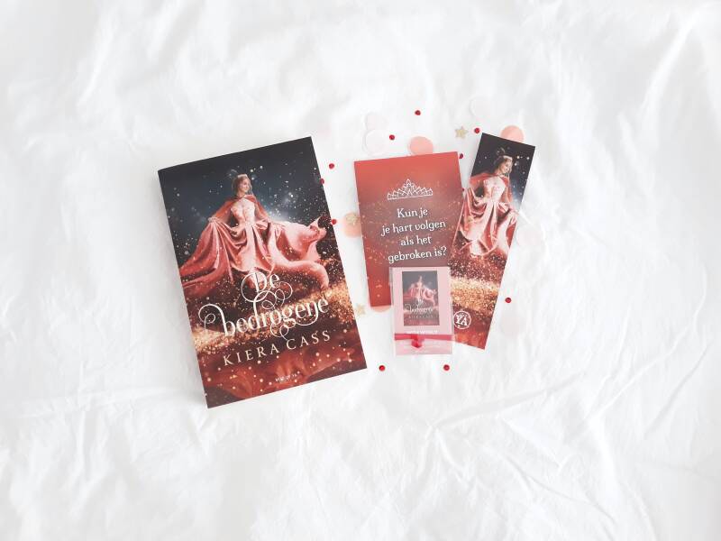 Op een wit kreukelig laken ligt het rode boek 'de bedrogene' met een paar bijpassende goodies; een rode quote-kaart, bijpassende boekenlegger en een kaartje met wensarmbandje gemaakt door @applepiepieces. Ook ligt er wat rode en roze confetti om de foto w