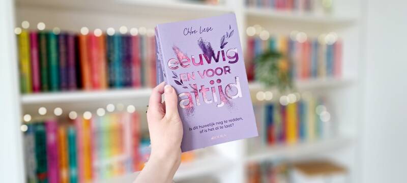 Een witte hand houdt 'Eeuwig en voor altijd' van CHloe Liese omhoog voor een kleurrijke wazige boekenkast met lichtjes. Foto bij recensie door turnthepages.nl | Disabled bookstagrammer en boekblogger.