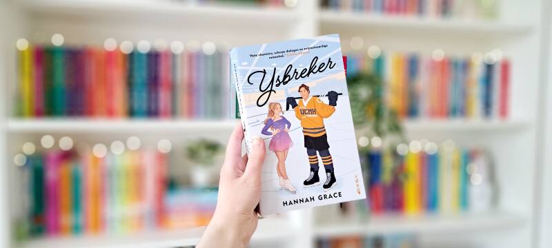 Een witte hand houdt 'IJsbreker' van Hannah Grace voor een kleurrijke wazige boekenkast met lichtjes. Foto bij recensie van turnthepages.nl Rolstoelgebruiker en disabled bookstagrammer en blogger.