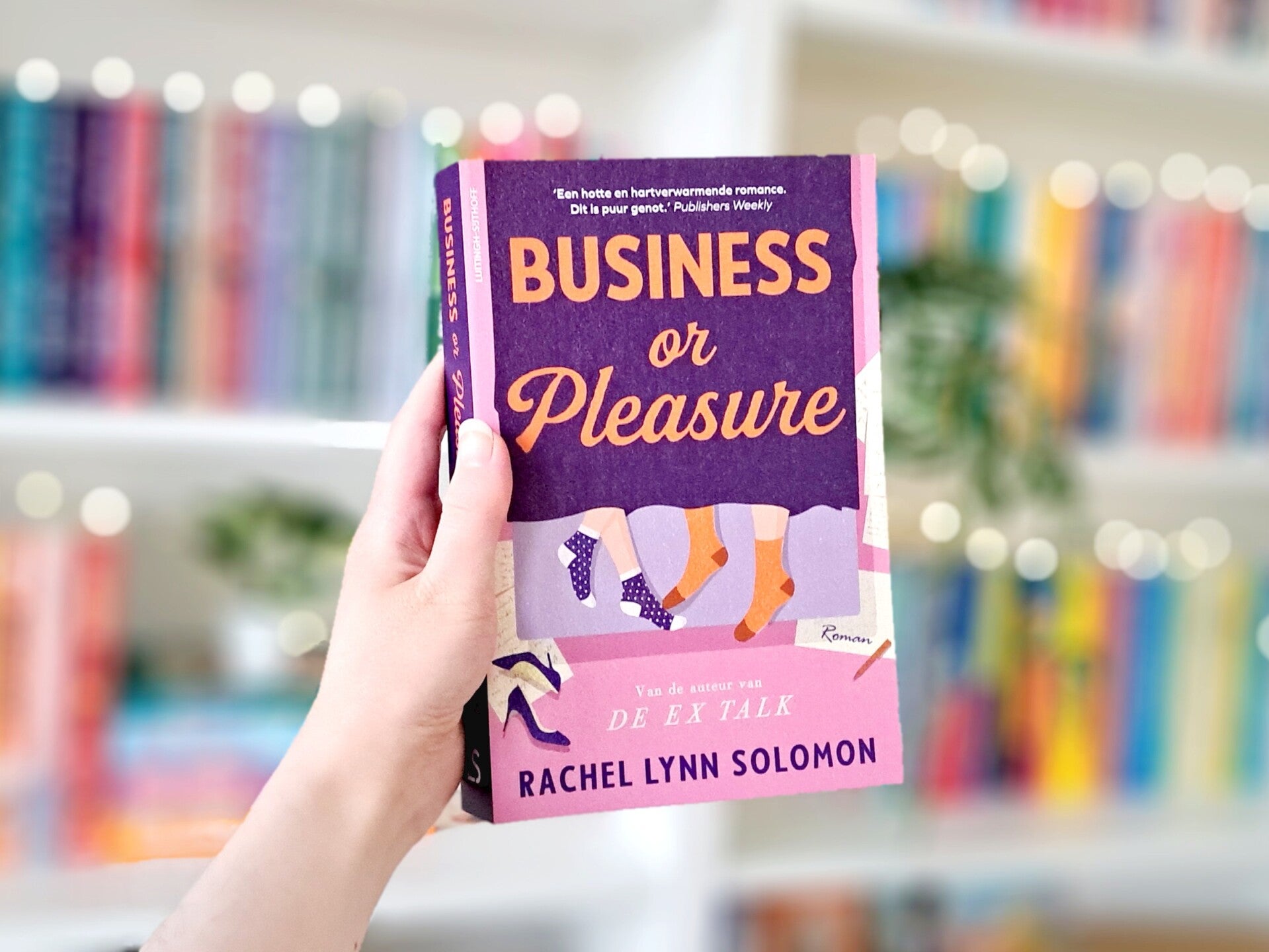 Een witte hand houdt de Nederlandse editie van 'Business or pleasure' van Rachel Lynn Solomon omhoog voor een kleurrijke wazige boekenkast met lichtjes. Foto bij recensie door turnthepages.nl | Disabled bookstagrammer en boekblogger met ADHD.