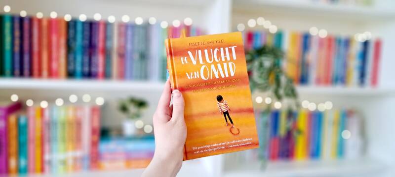 Een witte hand hodut 'de vlucht van Omid'geschreven door Lysette van Geel omhoog tegen een wazige boekenkast met lichtjes. Foto bij recensie door turnthepages.nl Rolstoelgebruiker en disabled bookstagrammer en blogger