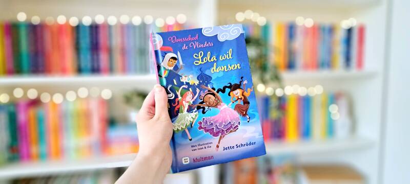 Een witte hand houdt 'Lola wil dansen' 'dansschool de vlinders deel 1'van Jette Schröder omhoog voor een kleurrijke wazige boekenkast met lichtjes. Foto bij recensie door turnthepages.nl Rolstoelgebruiker en disabled bookstagrammer en blogger.