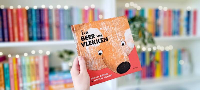 Een witte hand houdt 'Een beer met vlekken'van Monique Bodegom en Natascha Stenvert omhoog voor een kleurrijke wazige boekenkast met lichtjes. Foto bij recensie van turnthepages.nl rolstoelgebruiker en disabled bookstagrammer en blogger