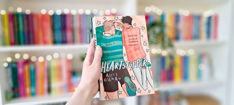 Een witte hand houdt 'Heartstopper #2 Nick en Charlie worden verliefd' van Alice Oseman omhoog voor een kleurrijke wazige boekenkast met lichtjes. Foto bij recensie door turnthepages.nl Disabled bookstagrammer en queer blogger.