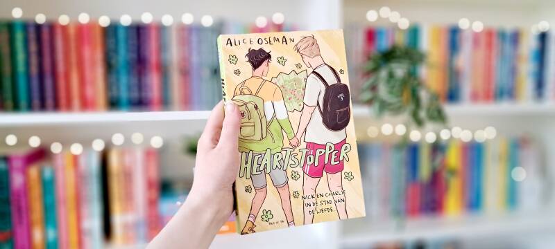 Een witte hand houdt 'Heartstopper #3 Nick en Charlie in de stad van de liefde' van Alice Oseman omhoog voor een kleurrijke wazige boekenkast met lichtjes. Foto bij recensie door turnthepages.nl Disabled bookstagrammer en queer blogger.